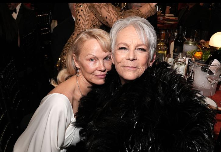 Pamela Anderson and Jamie Lee Curtis are seated at a table. Anderson rests her chin on Curtis's shoulder.