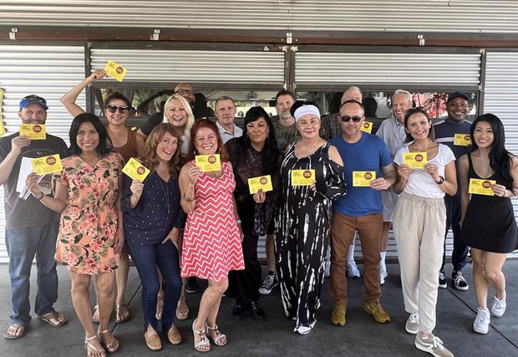 Seventeen members pose together holding bright yellow postcards. They are a diverse, group of men and women in an outdoor casual setting.