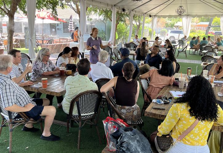A woman in a light purple dress is standing near the center, gesturing as she speaks to the attendees. The participants are seated around wooden tables, many with drinks in front of them. Papers and materials are spread out on the tables. The atmosphere is casual and has a collaborative and focused vibe.