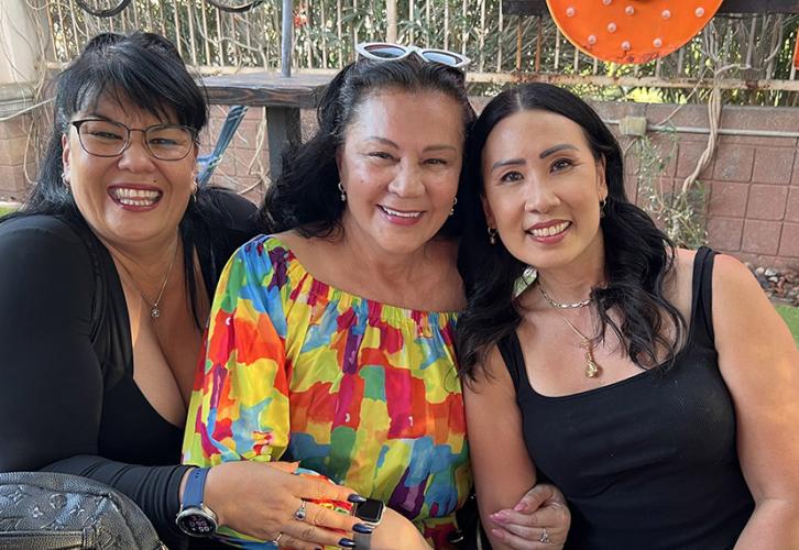 This image features three women outside seated closely together smiling. The woman on the left has dark hair, glasses, and is wearing a black top. The woman in the middle is wearing a colorful blouse. The woman on the right has long dark hair and is wearing a black tank top.
