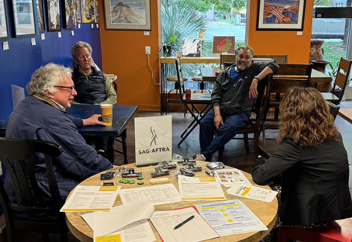 Four people seated in a cozy indoor cafe setting gather around two tables and engage in conversation. The nearest table is covered with SAG-AFTRA materials, including a sign with the organization’s logo, informational flyers, brochures and small promotional items. The background includes blue and orange walls adorned with framed artwork. The individuals are dressed casually, with one person holding a coffee cup.