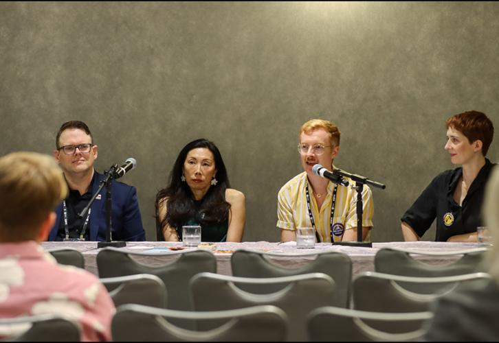 From left, Crabtree-Ireland, Long, Bowen and Elmaleh are seated at a table with microphones and two glasses of water. Bown is captured mid-speech and Elmaleh turns toward him. Crabtree-Ireland and Long stare forward listening intently. In the foreground are chairs and a person wearing a pink shirt with white flowers, far left.