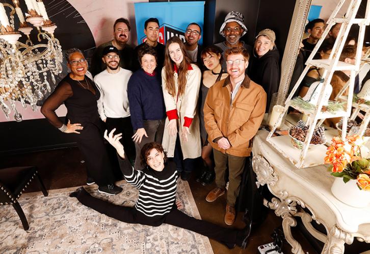 A diverse group of 12 individuals in a restaurant setting. One person in the front of the photo poses in a split on the floor with their right hand raised.