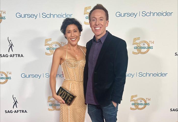 A woman and a man stand together on the red carpet. The woman wears a gold dress and black heels and holds a purse that says “Vote” in all-cap lettering. The man wears a blazer, a dark dress shirt and jeans. Behind them is a white step-and-repeat backdrop with the 50th Groundlings Anniversary, SAG-AFTRA and Gursey-Schneider logos
