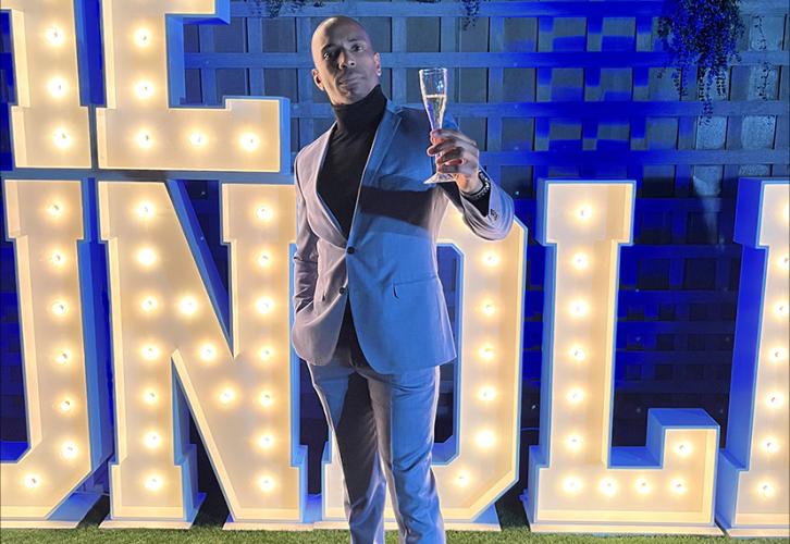 Robinson is outside in a grassy area. He wears a suit and a black turtleneck shirt. In his left hand is a glass of champagne, which he raises slightly. Partially displayed behind him are lighted letters in all caps.