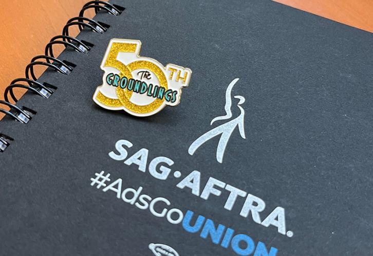 An image of a “The Groundlings 50th Anniversary” pin resting on a SAG-AFTRA #AdsGoUnion notebook. The pin has gold lettering, except for “Groundlings” which is green. The notebook is black and features the SAG-AFTRA and “#AdsGoUnion” logos. Except for “Union,” which is in all-cap blue lettering, the text and logo are white. 