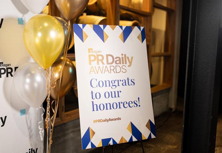Una imagen atmosférica de la alfombra roja. En un lugar destacado se encuentra un cartel con el logotipo de los PR Daily Awards en letras doradas y la frase “¡Felicitaciones a nuestros homenajeados!” en azul. Junto al cartel hay un manojo de globos dorados, blancos y amarillos y el fondo blanco con el logo de PR Daily.