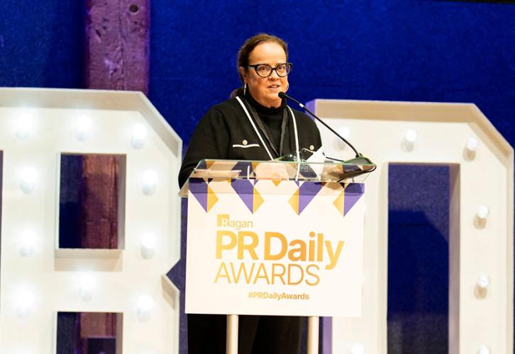 Greenwalt se encuentra en el escenario detrás de un podio con el logotipo de los PR Daily Awards. Mira hacia la multitud. Detrás de ella hay grandes letras “R” y “D” y un proyector de pantalla.