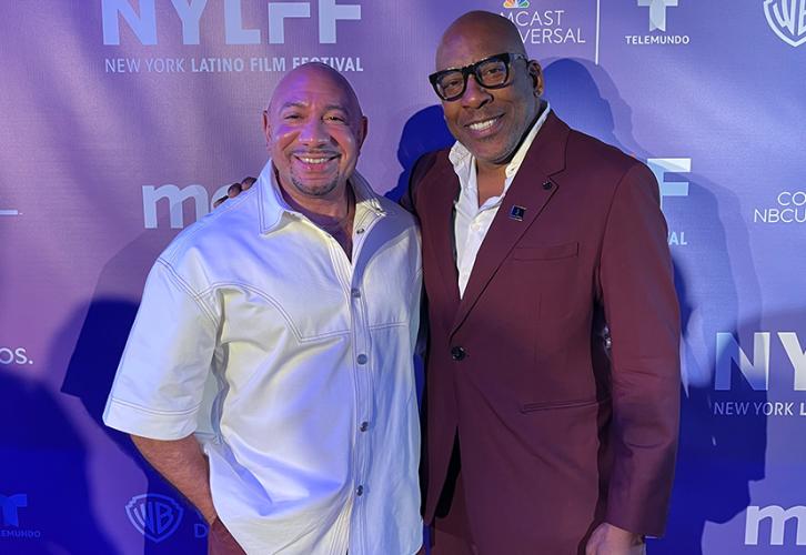 Chinchilla and Knight stand in front of a blue backdrop with logos for the New York Latino Film Festival, Warner Bros. Discovery, and Comcast NBCUniversal/Telemundo in white. Chinchilla wears a white shirt and brown pants; Knight wears a dark suit with a SAG-AFTRA button on the lapel and a white dress shirt. The lighting in the photo is a cool blue.