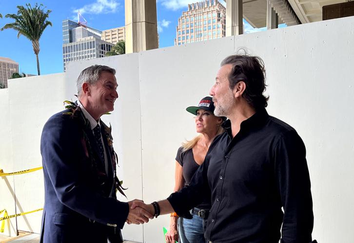 Dos personas se dan la mano mientras otra persona observa. La persona de la izquierda lleva un collar de hojas de ti.