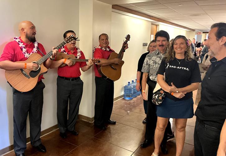 Tres personas vestidas de rojo y con leis están de pie en un salón tocando guitarras, mientras un pequeño grupo se detiene a mirar.
