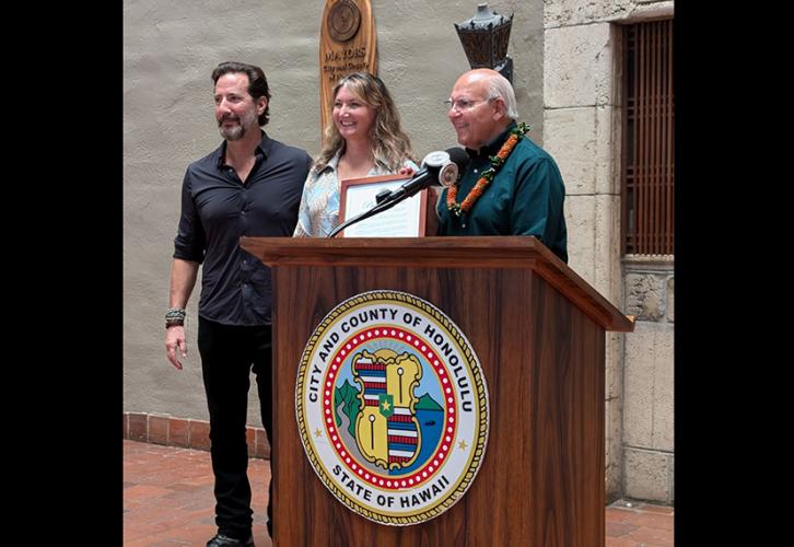 The Mayor of Honolulu, an white, older, avuncular gentleman in a dark green oxford shirt stands at podium on which the seal of the city and county of Honolulu is affixed, as he presents an official framed proclamation to the Hawaii Local President, Andy Sikkink, a middle aged white woman with wavy blonde hair, wearing a floral shirt, both are happily grinning. Standing to the left of her, National Board and Hawaii Local Board member Henry Ian Cusick, a fit middle age peruvian-scotsman.