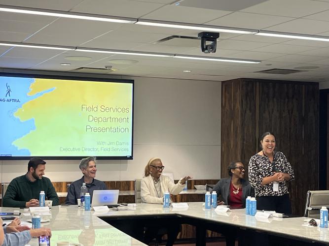 Four people are seated at a large table, with a fifth person standing to the far right. Behind them on the screen is a slide that reads “Field Services Department Presentation with Jim Damis, Executive Director, Field Services” in black text on a yellow and white background.