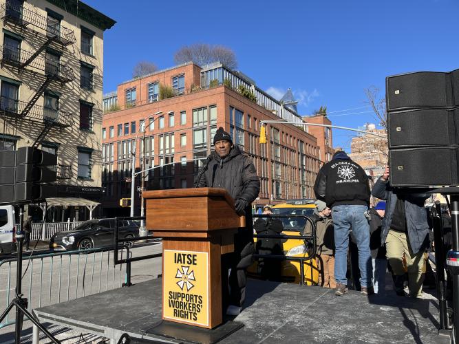 A man in a black winter coat and black beanie stands in front of a wooden podium with a yellow banner which reads “IATSE Supports Workers’ Rights” in black font.