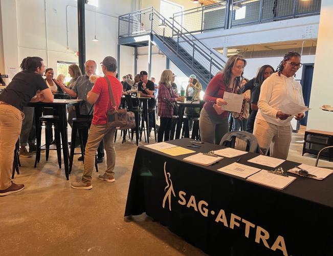 A large, diverse group of people interact with each other inside an industrial-like setting. Some stand around a table; others stand in a line holding papers they receive from a table. The table itself has a black SAG-AFTRA table cloth and various piles of paper.