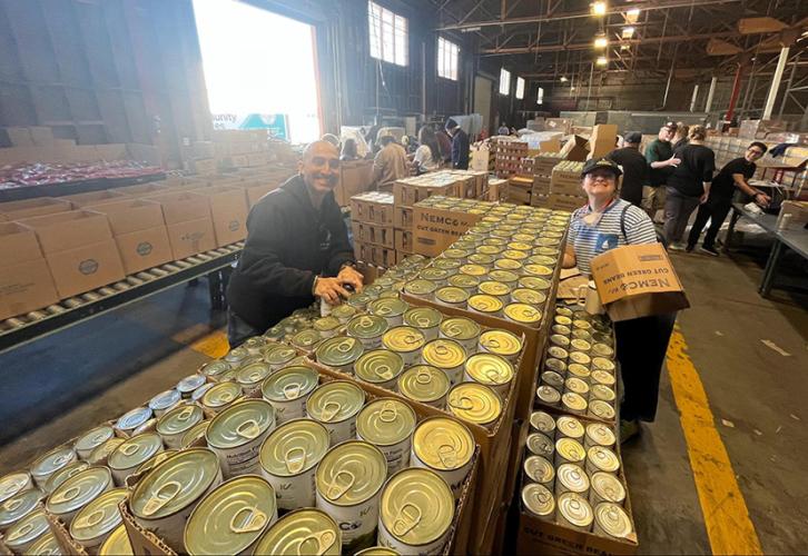 Two members with rows of canned goods in brown boxes in a warehouse.