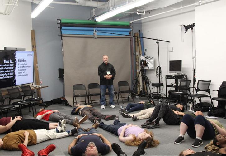 A group of individuals lay on the ground with one individual standing at the front of a classroom with a backdrop behind them.