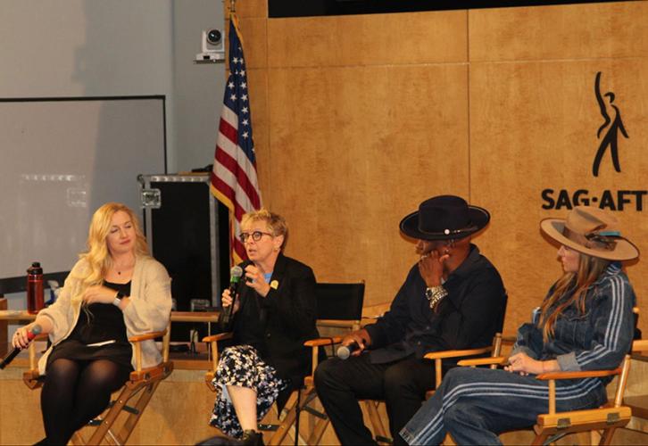 Four individuals sit in directors chairs in front of a wood-paneled wall with the SAG-AFTRA logo — a black figure with an upraised right arm with “SAG-AFTRA” underneath. There is an American flag in the background.
