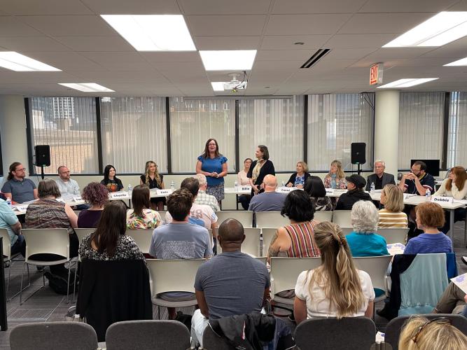 A wide shot of an office or conference room taken from the back of the room. Two women stand in front of a seated audience. One woman wearing a blue shirt and patterned skirt directly addresses the crowd. Behind her and seated at a long table are about nine or 10 people. 
