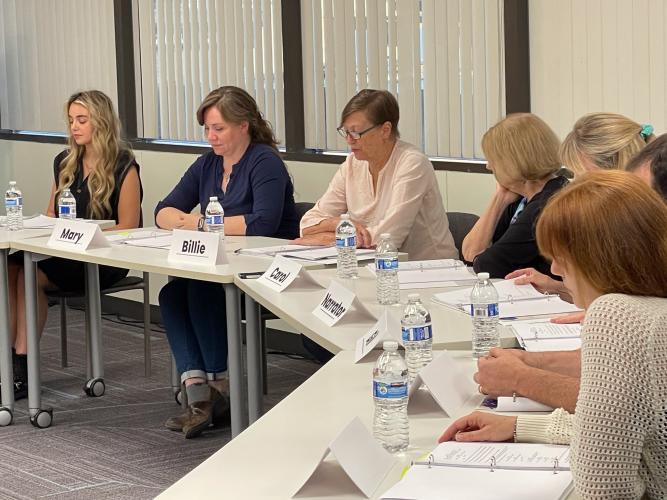 Seven individuals are seated at a long table. Three women are seen reading their scripts; the rest appear in a side profile. The tables are covered with papers and water bottles; positioned in the front are paper name cards bearing the names of characters.