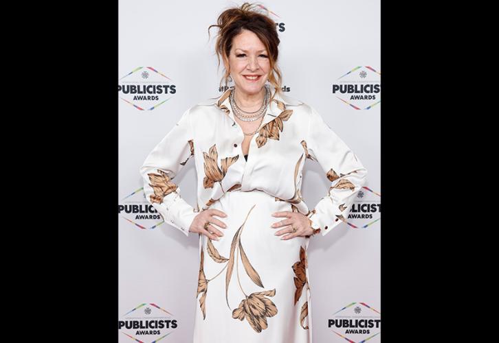 Fisher está en la alfombra roja. Luce un traje blanco con un estampado de flores de tallo y está de pie con las manos en las caderas. Detrás de ella hay un telón blanco con el logo de los premios.
