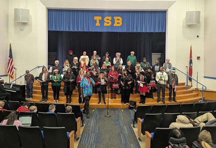 A wide shot of a large diverse group of singers arranged in rows onstage sing in front of a crowd. The group stands on bleacher steps. Above the group is a curtain with “TSB” in large gold lettering. The audience is visible in the foreground.