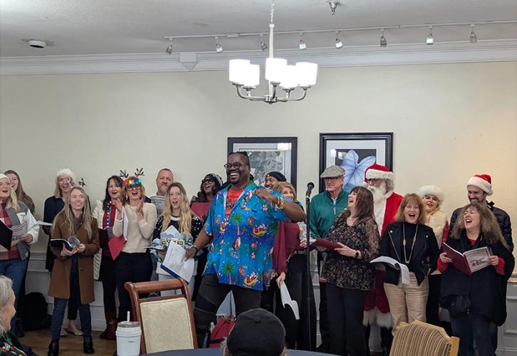 A group of singers arranged into two long rows sing in a small dining area. In the front is a tall man wearing a blue shirt patterned with images of Santa Claus and palm trees. In the foreground are some seated people and a small round dining table. 
