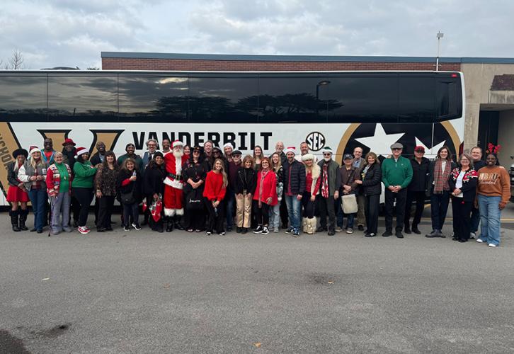 A large diverse group of people stand outside in a parking lot or loading dock area. All are dressed in holiday festive clothing: green and red; Santa Claus hats. One gentleman in front is dressed as Santa Claus. Behind the group is a large tour bus bearing Vanderbilt University’s school colors and logos. 