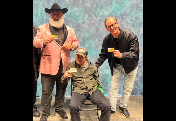Three individuals take a photo in front of a photo backdrop. Fox, center, is seated and wears a dark green dress shirt over a gray t-shirt, dark slacks and a baseball cap. They all hold a SAG-AFTRA membership card.