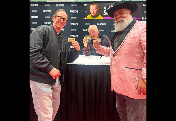 Three individuals take a photo in an exhibition hall. Shatner, center, is seated behind a table and wears a black vest over a dark purple dress shirt. Behind them is a black Fan Expo New Orleans step-and-repeat backdrop with a banner bearing Shatner’s name and an image of him as ‘Star Trek’ character Captain Kirk.