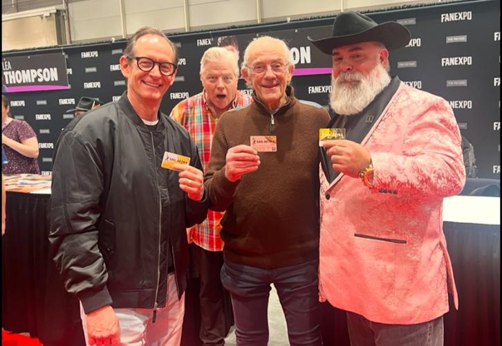 Four individuals take a photo in an exhibition hall. Lloyd wears a brown shirt and dark slacks. Each of them holds a SAG-AFTRA membership card. Behind the group is Wilson who makes a shocked expression toward the camera. There are also tables and a black Fan Expo New Orleans step-and-repeat backdrop.