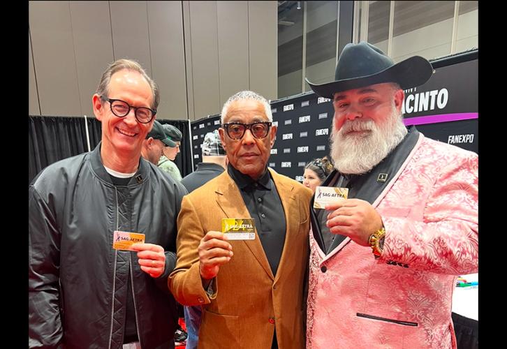 Three individuals take a photo in an exhibition hall. Esposito wears a gold blazer over a black dress shirt and slacks. Each of them holds a SAG-AFTRA membership card. Behind them is a black Fan Expo New Orleans step-and-repeat backdrop.