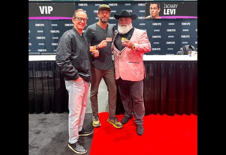 Three individuals take a photo in an exhibition hall. Levi wears a gray shirt, brown slacks, and green-and-black sneakers. Each of them holds a SAG-AFTRA membership card. There are also tables and a black Fan Expo New Orleans step-and-repeat backdrop with Levi’s name and photo of his character from ‘Shazam!’