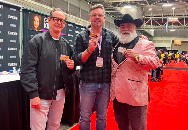 Three individuals take a photo in an exhibition hall. Welling wears a dark-colored flannel shirt and jeans. Each of them holds a SAG-AFTRA membership card. There are also tables and a black Fan Expo New Orleans step-and-repeat backdrop.