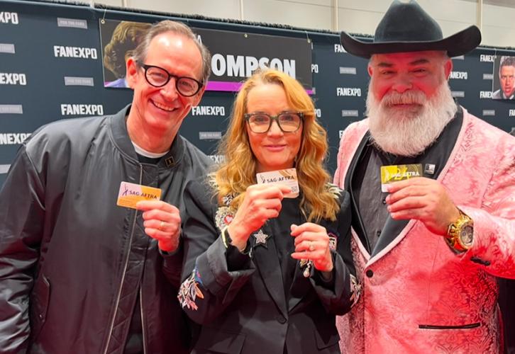 Three individuals take a photo in an exhibition hall. Thompson wears a black blazer jacket with a silver brooch over a black shirt. Each of them holds a SAG-AFTRA membership card in their hands. Behind them is a black Fan Expo New Orleans step-and-repeat backdrop with a banner featuring Thompson’s name and photo.