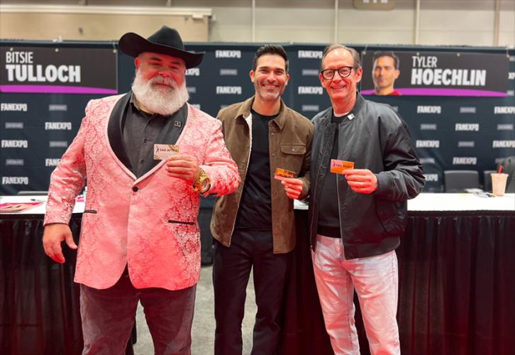 Three individuals take a photo in an exhibition hall. Hoechlin wears a brown jacket over a black shirt and pants. Each of them holds a SAG-AFTRA membership card in their hands. There are also tables and a black Fan Expo New Orleans step-and-repeat backdrop.