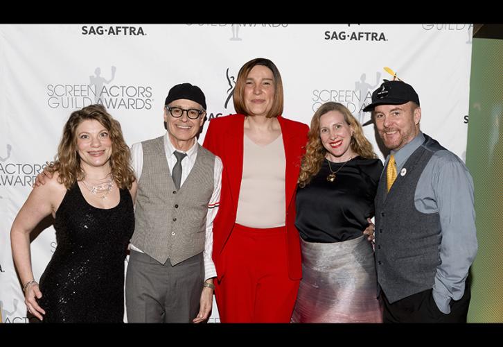 Five individuals take a photo on the red carpet. All are dressed semi-formally; the second and fifth individuals wear hats. Behind the group is a white step-and-repeat backdrop with the SAG-AFTRA and SAG Awards logos.