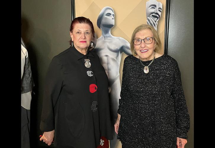 Two individuals take a photo together. Both wear all-black ensembles. Behind them is a banner of the SAG Awards statuette.  