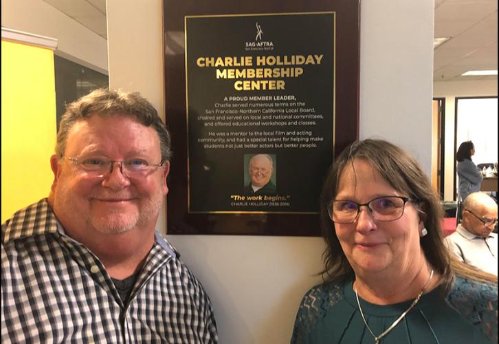 Two people with glasses stand on either side of a wooden plaque with a photo and an inscription reading “Charlie Holliday Membership Center.”