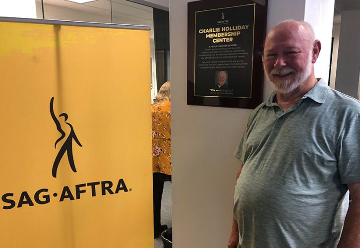 A person with a beard, smiling, standing next to a wooden photo plaque hung on a white column stating “Charlie Holliday Membership Center.” To the left is a yellow standing SAG-AFTRA banner.