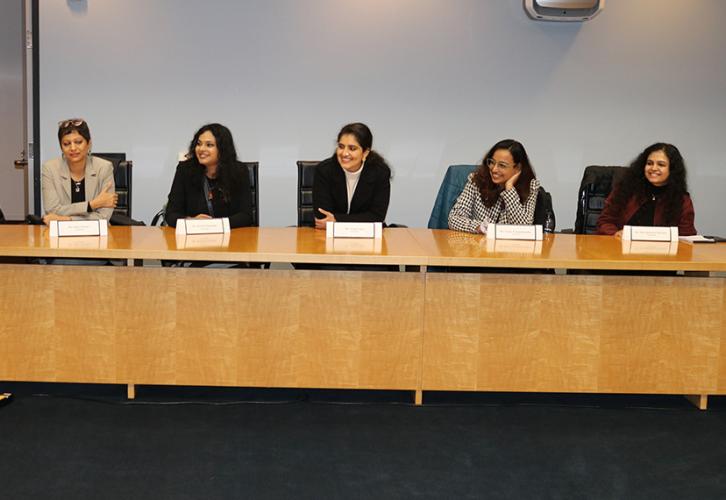Five individuals are seated at a table within a nondescript conference room. All are turned slightly to their right.