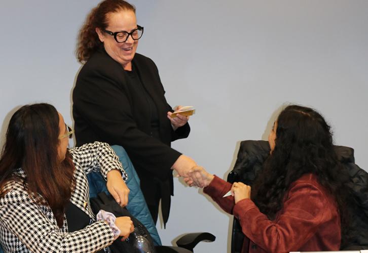 Three people in the photo, one standing and two seated at a table. The standing individual shakes hands with the person seated on the left. The person seated on the right looks on.