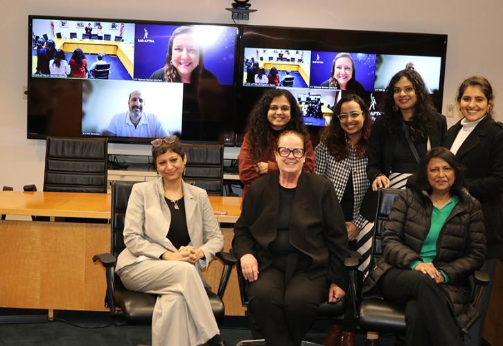 A group shot of seven individuals in front of a large flatscreen. Three people in the group are seated; four are standing. On the flatscreen are squares as part of a virtual meeting. Two individuals are visible.