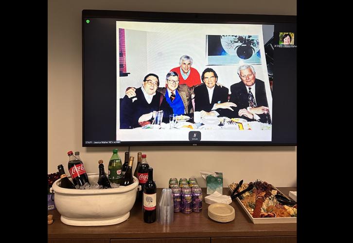 Various sodas and wines are arranged in a wine bucket with ice beside an assortment of seltzers, plates and napkins, and a cheeseboard on a counter in the SAG-AFTRA New England Local Boardroom. A photo of John Henning and four of his former colleagues is projected on the screen behind the cabinet.