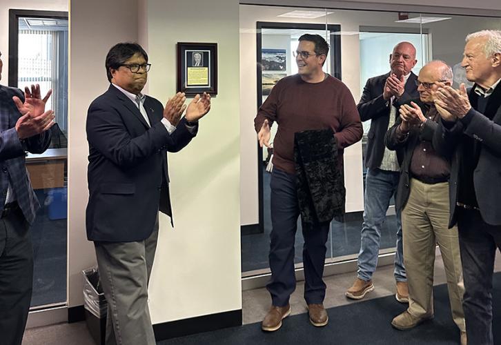 A group of four broadcasters and one of John Henning’s sons clap after taking off the black tarp and revealing the plaque honoring his legacy. His other son holds the tarp while smiling.