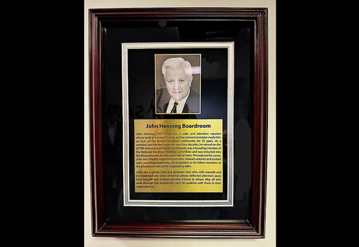 A brown, wooden frame borders a portrait of John Henning and written speech about his accomplishments displayed on a gold background and framed within a black backdrop. The title of the plaque, spelled out below his portrait and above the speech, reads “John Henning Boardroom.”