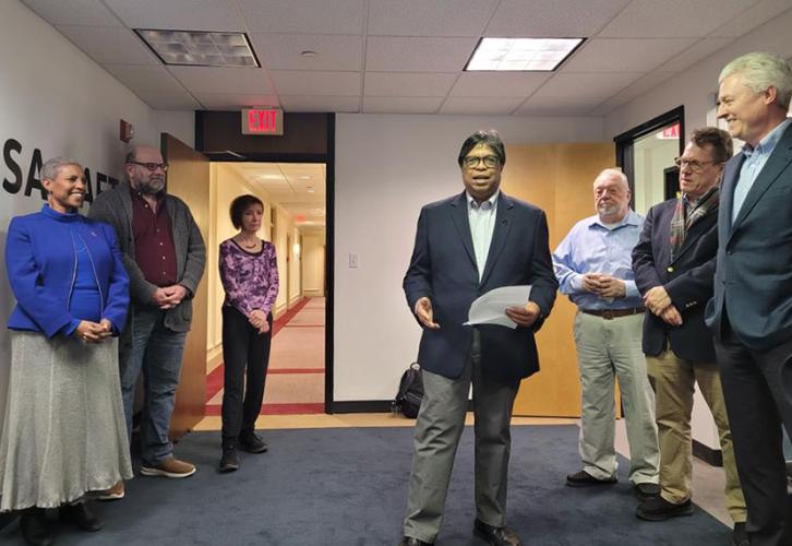 Six former colleagues of broadcaster John Henning stand around SAG-AFTRA New England board member Vic Ramos as he presents a speech about the career of John Henning prior to revealing the plaque dedicated to his legacy.