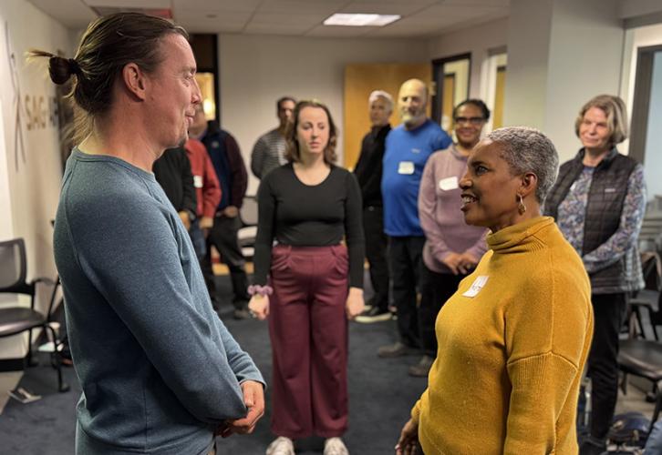 Two SAG-AFTRA members make eye contact – one in a yellow sweater and one in a blue long-sleeved shirt – as part of an acting exercise. Several members and one instructor watch the demonstration in the background. 