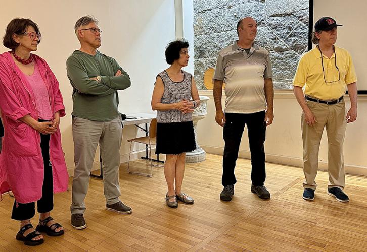 Five people stand in a semi-circle as part of an interactive acting exercise. The first is in a long pink shawl, chunky pink jewelry and red glasses. The second person is in gray pants and a green long-sleeve shirt. The third person is wearing a black skirt and a sleeveless black-and-white top. The fourth person is wearing blank pants and a striped collared T-shirt. The fifth person is wearing khaki pants, a belt, a yellow-collared T-shirt, a Red Sox hat, and has reading glasses hanging around their neck.