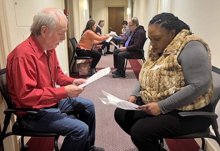 Pairs of SAG-AFTRA New England members sit in chairs; one pair in the foreground consists of a member in a red button-down and jeans and a member in a brown vest and black pants. Two more groups of two can be seen in the background further down the hallway.